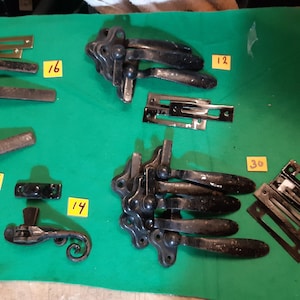 May include: An assortment of antique black metal hardware components, including hinges, latches, and mounting plates, are arranged on a green surface. Some pieces have brass accents. Yellow tags with numbers are placed near the hardware.