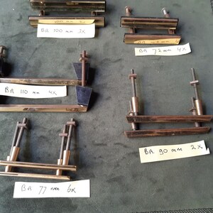 Op de afbeelding: Vijf sets metalen ladehandgrepen met draadeinden en moeren, voorzien van handgeschreven briefjes: BR 100 mm 3x, BR 72 mm 4x, BR 110 mm 4x, BR 90 mm 2x en BR 77 mm 6x. Het beslag heeft een gepolijste afwerking en bevat zwarte montageblokken.