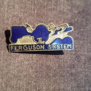 Op de afbeelding: Een rechthoekige metalen speld met een diepblauwe emaille inleg. Het ontwerp bevat een gestileerd silhouet van een tractor en de tekst The FERGUSON SYSTEM in verhoogde zilverkleurige letters. De speld heeft een metalen rand.