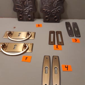 Peut inclure: Un assortiment de composants de quincaillerie en laiton antique, comprenant des poignées de tiroir, des cache-trous de serrure et des plaques arrière décoratives. La quincaillerie présente un mélange de motifs ornés et simples, avec une patine chaude et vieillie.