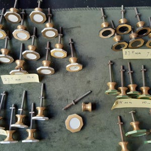 Peut inclure: Une collection de boutons en m&eacute;tal dor&eacute; avec des t&ecirc;tes octogonales et des centres circulaires blancs ou sombres, chacun dot&eacute; d'une longue tige filet&eacute;e. Des notes adh&eacute;sives jaunes portent des inscriptions manuscrites, dont 27,5 mm et 24 mm.