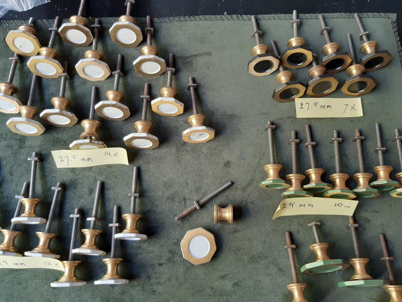 Peut inclure: Une collection de boutons en m&eacute;tal dor&eacute; avec des t&ecirc;tes octogonales et des centres circulaires blancs ou sombres, chacun dot&eacute; d'une longue tige filet&eacute;e. Des notes adh&eacute;sives jaunes portent des inscriptions manuscrites, dont 27,5 mm et 24 mm.
