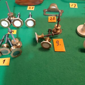 Op de afbeelding: Een verzameling antieke meubelbeslag, waaronder knoppen en een handgreep, gepresenteerd op een groene ondergrond. De knoppen hebben verschillende vormen en kleuren, sommige met witte of groene accenten. Kleine oranje labels met nummers identificeren elk stuk.