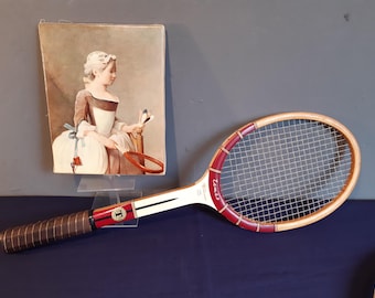 May include: A vintage wooden tennis racket with a brown leather grip, red and white accents, and the word "Temzo" printed on the frame. Beside it is a rectangular print depicting a person in historical attire holding a similar tennis racket.