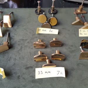 Könnte beinhalten: Eine Sammlung goldfarbener Metall-Möbelknöpfe in verschiedenen Formen, darunter trapezförmige, kreisförmige und rechteckige Designs. Mehrere Knöpfe sind mit gelben Klebezetteln versehen, die handschriftliche Notizen wie 30 mm 6x, H 22 mm BA 40 mm 12x und 33 mm 7x tragen und die jeweiligen Maße und Mengen angeben.