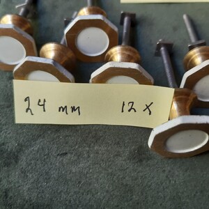 Peut inclure: Un ensemble de douze boutons de placard avec un cadre octogonal en laiton et un insert circulaire blanc au centre. Chaque bouton poss&egrave;de une tige filet&eacute;e en m&eacute;tal &agrave; l'arri&egrave;re. Une note en papier jaune porte l'inscription 24 mm 12 x.