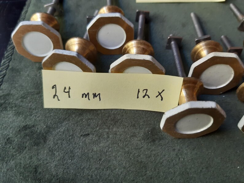 Peut inclure: Un ensemble de douze boutons de placard avec un cadre octogonal en laiton et un insert circulaire blanc au centre. Chaque bouton poss&egrave;de une tige filet&eacute;e en m&eacute;tal &agrave; l'arri&egrave;re. Une note en papier jaune porte l'inscription 24 mm 12 x.
