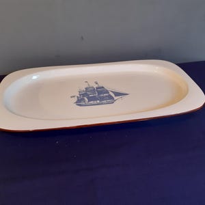 Op de afbeelding: Een witte, rechthoekige serveerschaal met een blauwe zeilbootillustratie in het midden. De schaal heeft een bruine rand en staat tegen een blauwe achtergrond. Dit gerecht met nautisch thema is ideaal voor het serveren van hapjes of desserts.