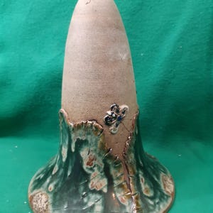 Op de afbeelding: Een handgemaakte keramische flesstopper met een uniek ontwerp. De stopper heeft een lichtbruine, conische bovenkant en een uitlopende basis met een groen en bruin geglazuurde afwerking. Een kleine, decoratieve vlinder is aan de basis bevestigd.