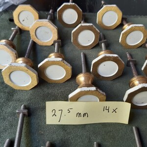 Peut inclure: Un ensemble de quatorze boutons de placard octogonaux avec un centre circulaire blanc entour&eacute; d'une bordure dor&eacute;e. Chaque bouton comprend une tige filet&eacute;e en m&eacute;tal et un &eacute;crou. Une note jaune affiche le texte 27.5 mm 14x, indiquant la taille.