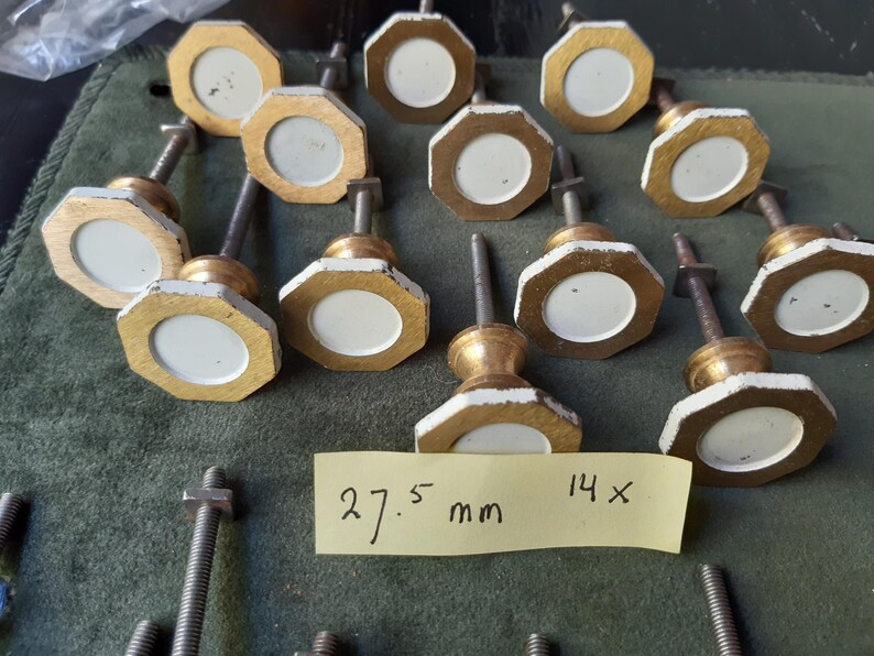 Peut inclure: Un ensemble de quatorze boutons de placard octogonaux avec un centre circulaire blanc entour&eacute; d'une bordure dor&eacute;e. Chaque bouton comprend une tige filet&eacute;e en m&eacute;tal et un &eacute;crou. Une note jaune affiche le texte 27.5 mm 14x, indiquant la taille.