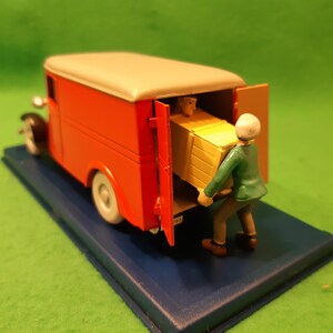 以下が含まれることがあります： クリーム色の屋根と白いタイヤが付いた、オレンジ色のミニチュア配達トラック。緑色のジャケットを着た人物が、黄色の箱をトラックに積み込んでいる。トラックは青い台座の上にあり、緑色の背景に配置されている。