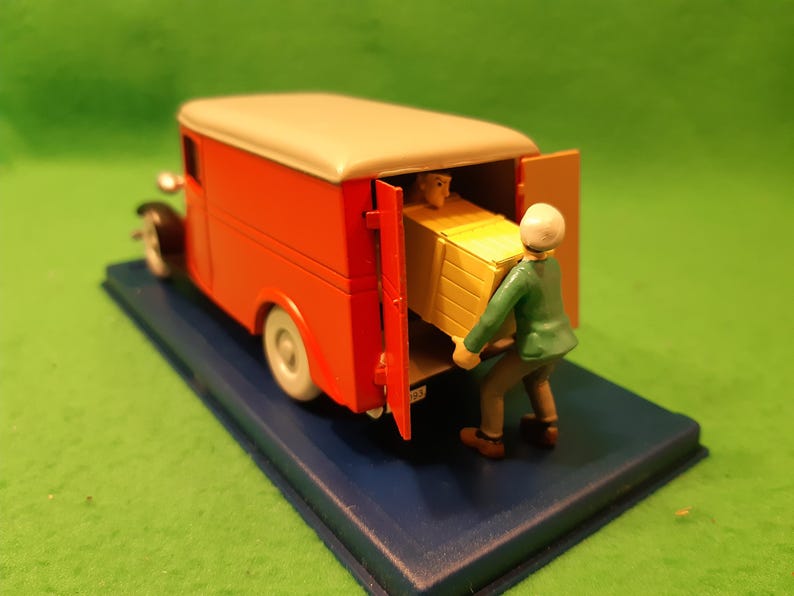 以下が含まれることがあります： クリーム色の屋根と白いタイヤが付いた、オレンジ色のミニチュア配達トラック。緑色のジャケットを着た人物が、黄色の箱をトラックに積み込んでいる。トラックは青い台座の上にあり、緑色の背景に配置されている。