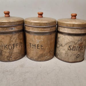 May include: Three cylindrical metal canisters with a weathered, marbled beige finish and dark metal bands. Each canister features a small wooden knob on the lid and black lettering reading KOFFIE, THEE, and SUIKER on the front of the containers.