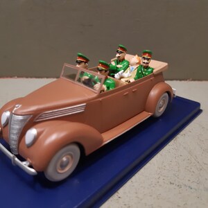 May include: A brown vintage toy car with four figures in green uniforms and hats. The car has a tan convertible top and is on a blue base. The car has a silver front bumper and headlights.