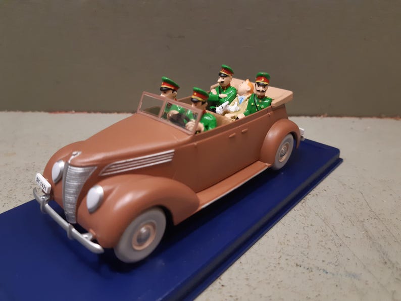 May include: A brown vintage toy car with four figures in green uniforms and hats. The car has a tan convertible top and is on a blue base. The car has a silver front bumper and headlights.