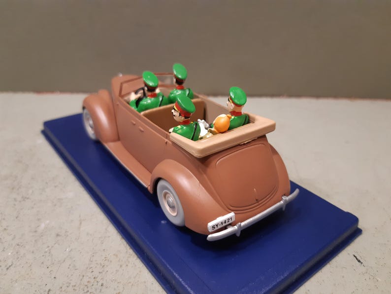 May include: A brown vintage convertible toy car with four figures in green uniforms and hats. The car sits on a blue base. The car has white tires and a license plate that reads "SY-1421".