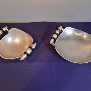 May include: Two silver-toned metal serving bowls with handles. Each bowl has a rounded, asymmetrical shape and is equipped with two handles. The handles are silver with black accents. The bowls are displayed on a dark blue surface.
