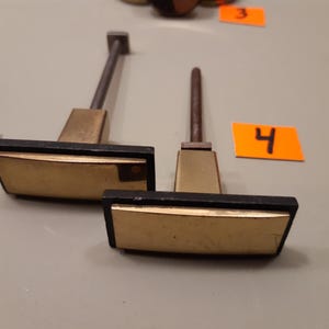 Może przedstawiać: Dwa uchwyty do szafek w stylu vintage z prostokątnymi, złotymi powierzchniami i czarnym wykończeniem. Uchwyty mają podstawę w kolorze mosiądzu i ciemny metalowy trzpień. Numer "4" znajduje się na pomarańczowej etykiecie.