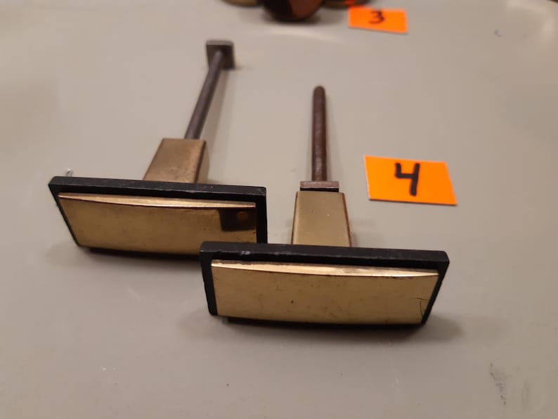 Może przedstawiać: Dwa uchwyty do szafek w stylu vintage z prostokątnymi, złotymi powierzchniami i czarnym wykończeniem. Uchwyty mają podstawę w kolorze mosiądzu i ciemny metalowy trzpień. Numer "4" znajduje się na pomarańczowej etykiecie.