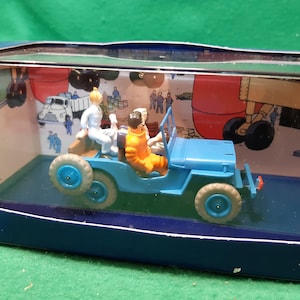 Puede incluir: Un jeep de juguete azul con dos figuras dentro, presentado en una caja expositora. El jeep tiene ruedas azules y una carrocer&iacute;a azul. Las figuras visten trajes blancos y naranjas. El fondo de la caja presenta una escena de dibujos animados.
