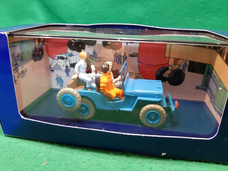Puede incluir: Un jeep de juguete azul con dos figuras dentro, presentado en una caja expositora. El jeep tiene ruedas azules y una carrocer&iacute;a azul. Las figuras visten trajes blancos y naranjas. El fondo de la caja presenta una escena de dibujos animados.