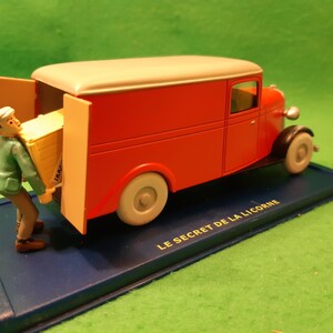 以下が含まれることがあります： 赤いクリーム色のヴィンテージ配送バンモデル。ドアが開いており、フィギュアが黄色の箱を積み込んでいる。モデルは「LE SECRET DE LA LICORNE」の文字が入った青いベースの上に置かれている。