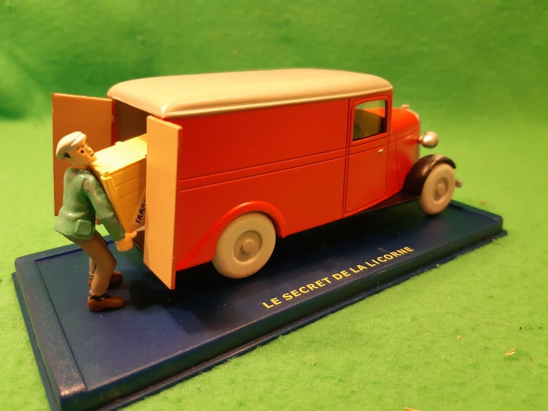 以下が含まれることがあります： 赤いクリーム色のヴィンテージ配送バンモデル。ドアが開いており、フィギュアが黄色の箱を積み込んでいる。モデルは「LE SECRET DE LA LICORNE」の文字が入った青いベースの上に置かれている。