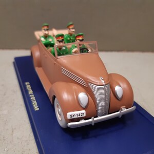 May include: A brown vintage convertible toy car with four figures in green uniforms and hats. The car has white headlights, a silver bumper, and a license plate that reads "SY-1421". The car sits on a blue base.