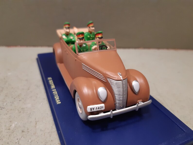 May include: A brown vintage convertible toy car with four figures in green uniforms and hats. The car has white headlights, a silver bumper, and a license plate that reads "SY-1421". The car sits on a blue base.