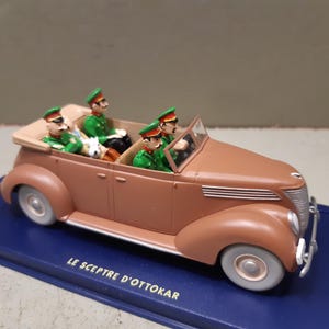 May include: A brown vintage toy car with four figures in green uniforms and hats. The car has white tires and a blue base with the text "LE SCEPTRE D'OTTOKAR". The car is a classic model, likely from the mid-20th century.