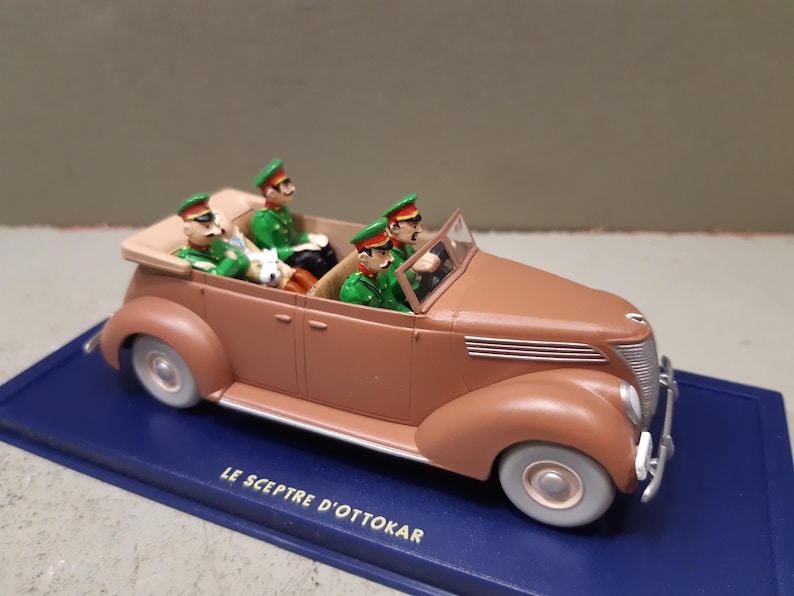 May include: A brown vintage toy car with four figures in green uniforms and hats. The car has white tires and a blue base with the text "LE SCEPTRE D'OTTOKAR". The car is a classic model, likely from the mid-20th century.