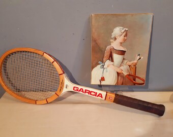 May include: A vintage wooden tennis racket with the brand name "GARCIA" in red lettering. The racket has a brown grip and a light-colored frame. A framed painting of a woman in a period dress is in the background.