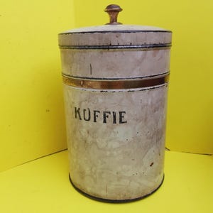 Può includere: Latta da caffè cilindrica vintage con coperchio e pomello in legno. La latta è bianco sporco con dettagli neri e dorati. La parola "KOFFIE" è stampata in nero. Un classico contenitore da cucina.