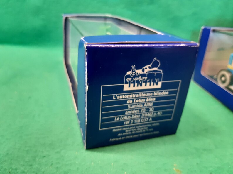K&ouml;nnte beinhalten: Blaue Box mit Tintin-Logo und franz&ouml;sischem Text. Der Text lautet "L'automitrailleuse blindee du Lotus bleu" und "Sumida ARM ann&eacute;es 20-30". Die Box befindet sich auf einer gr&uuml;nen Oberfl&auml;che.