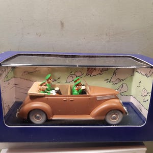 May include: A brown vintage convertible toy car with two figures in green uniforms and hats. The car is displayed in a blue box with a clear plastic window, set against a backdrop of a cartoon landscape.