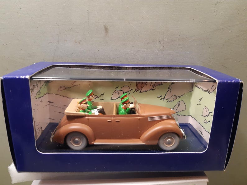 May include: A brown vintage convertible toy car with two figures in green uniforms and hats. The car is displayed in a blue box with a clear plastic window, set against a backdrop of a cartoon landscape.