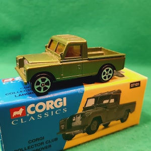 以下が含まれることがあります： 黒とシルバーのホイールが付いた、オリーブグリーンのダイキャスト製Corgi Classics Land Roverピックアップトラックのおもちゃが、オリジナルの青と黄色の箱の上に置かれています。箱にはCorgiのロゴとトラックのイラストが描かれています。