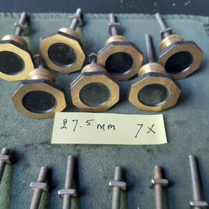 Peut inclure: Sept boutons octogonaux en laiton avec des centres circulaires noirs, chacun dot&eacute; d'une tige filet&eacute;e et d'un &eacute;crou en m&eacute;tal. Une note en papier jaune est plac&eacute;e au centre avec le texte manuscrit 27.5 mm 7x, indiquant le diam&egrave;tre des boutons.