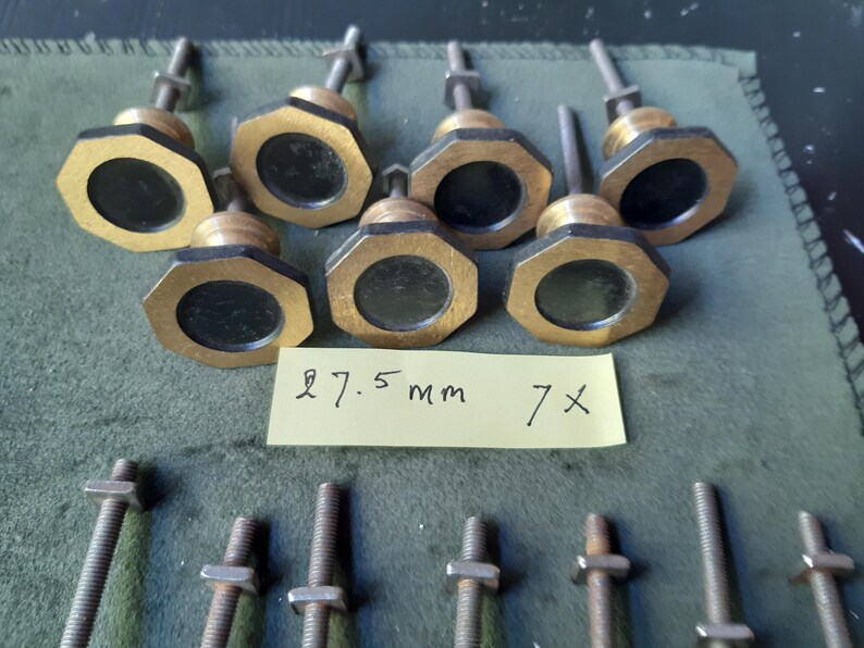 Peut inclure: Sept boutons octogonaux en laiton avec des centres circulaires noirs, chacun dot&eacute; d'une tige filet&eacute;e et d'un &eacute;crou en m&eacute;tal. Une note en papier jaune est plac&eacute;e au centre avec le texte manuscrit 27.5 mm 7x, indiquant le diam&egrave;tre des boutons.