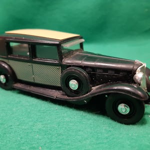 Könnte beinhalten: Ein schwarzes und beigefarbenes Spielzeugauto im Vintage-Stil. Es hat schwarze Reifen, einen silbernen Kühlergrill und ein beiges Dach. Die Seitenwände weisen ein strukturiertes Muster auf. Ein klassisches Modell.