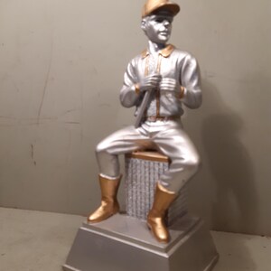 Könnte beinhalten: Eine silber- und goldfarbene Baseballspieler-Figur mit Kappe, Jacke und Hose, die auf einem quadratischen Block sitzt. Die Figur hält einen Baseballschläger vertikal vor der Brust und trägt hohe Stiefel, alles auf einem breiten silbernen Sockel.