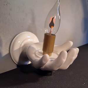 May include: A white ceramic wall sconce shaped like a hand holding a decorative light bulb. The light bulb is clear with a flame-shaped design and a gold-colored base. The sconce is mounted on a white circular base.