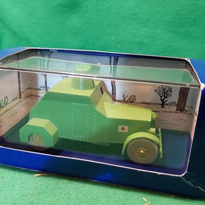 K&ouml;nnte beinhalten: Gr&uuml;nes Spielzeug-Panzermodell in einer blauen Box mit transparentem Deckel. Das Modell hat eine japanische Flagge. Die Box hat eine gedruckte Landschaftsszene im Inneren.