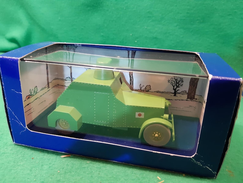 K&ouml;nnte beinhalten: Gr&uuml;nes Spielzeug-Panzermodell in einer blauen Box mit transparentem Deckel. Das Modell hat eine japanische Flagge. Die Box hat eine gedruckte Landschaftsszene im Inneren.