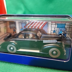 May include: A dark green vintage car model in a display box. The car has a black roof and silver-colored wheels. The box features a street scene backdrop with cartoon figures and buildings.