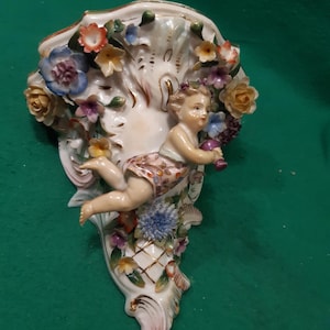 Puede incluir: Un bolsillo de pared de porcelana con una figura de querubín rodeada de flores coloridas. El querubín sostiene un racimo de uvas. El bolsillo tiene forma de concha con detalles dorados y un diseño de enrejado.