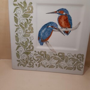 Op de afbeelding: Vierkante witte keramische plaat met een handgeschilderd ontwerp. Voorzien van twee blauwe en oranje ijsvogels die op een tak zitten. De plaat heeft een groen en wit bloemenrandontwerp. Een decoratief stuk voor in huis.