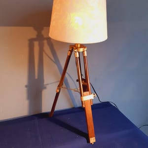 Può includere: Una lampada da tavolo con base a treppiede in legno marrone scuro con dettagli e finiture in ottone. La lampada è dotata di un paralume cilindrico in tessuto color bianco sporco che emette una luce calda, sostenuto da tre gambe in legno.