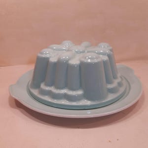Op de afbeelding: Een lichtblauwe keramische puddingvorm met een gegroefd ontwerp, op een bijpassende schotel. De vorm is cirkelvormig met verticale ribbels en een afgeronde bovenkant. De schotel heeft een licht verhoogde rand.